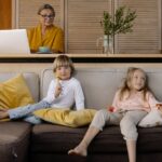 Starsza kobieta pracuje zdalnie na laptopie, wnuki bawią się na kanapie